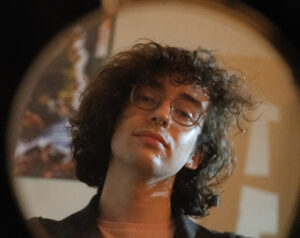 A headshot of a person with brown curly hair and glasses, with a blurred background.